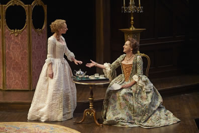 Dorinda is in an all-white floor-length dress, with corset and puffy sleeves, her blond hair done up, and she holds her hand out to the sitting Mrs. Sullen, in a green tapestry dress with gold corset and a neckless. Between them is a teaset on fine table, with, in the background, a calabra on a stand and a pink screen